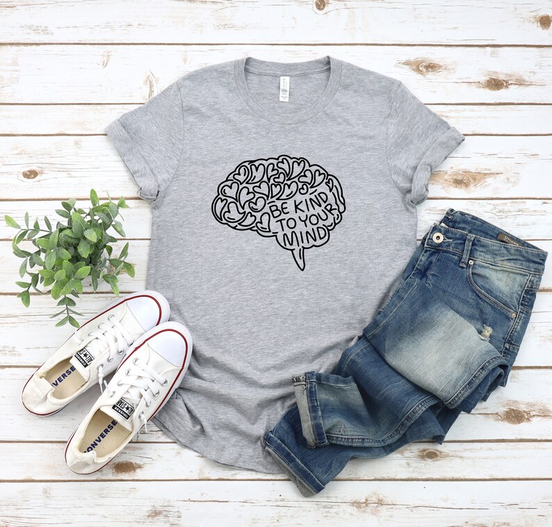 May include: Gray t-shirt with a black outline of a brain with the text "Be Kind To Your Mind" inside. The shirt is laid out on a white wood surface with a pair of white sneakers and blue jeans.