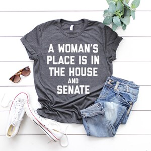 May include: A dark gray t-shirt with the text "A woman's place is in the house and senate" printed in white.