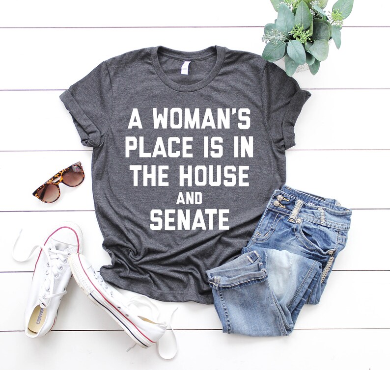 May include: A dark gray t-shirt with the text "A woman's place is in the house and senate" printed in white.