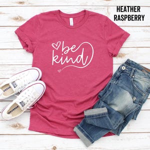 May include: A heather raspberry t-shirt with the text "be kind" in white lettering with a heart design. The shirt is laid flat on a wooden surface with a pair of white sneakers and blue jeans.
