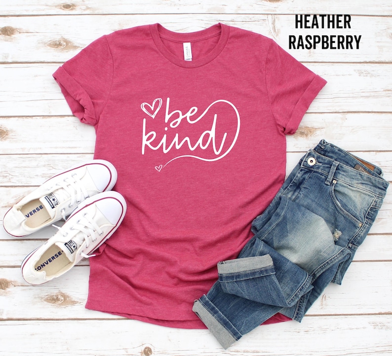 May include: A heather raspberry t-shirt with the text "be kind" in white lettering with a heart design. The shirt is laid flat on a wooden surface with a pair of white sneakers and blue jeans.