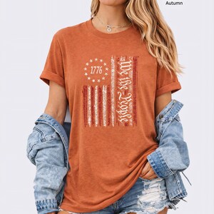 May include: Heather autumn-colored t-shirt with a distressed American flag design. The flag features red and white stripes, a circle of stars with the number "1776", and the words "We the People" in white.