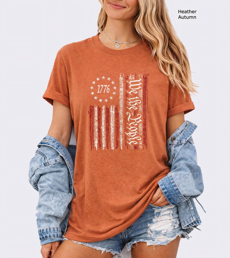 May include: Heather autumn-colored t-shirt with a distressed American flag design. The flag features red and white stripes, a circle of stars with the number "1776", and the words "We the People" in white.