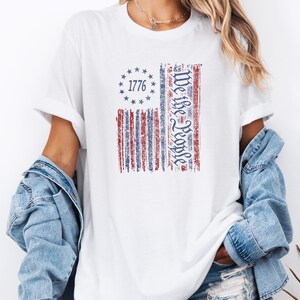 May include: White t-shirt with a distressed American flag design. The flag features red and blue stripes, a circle of stars, and the text "1776" and "We the People". The shirt is worn with a denim jacket.