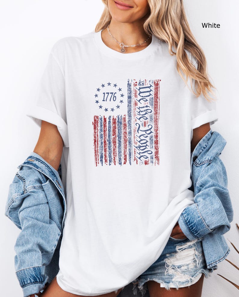 May include: White t-shirt with a distressed American flag design. The flag features red and blue stripes, a circle of stars, and the text "1776" and "We the People". The shirt is worn with a denim jacket.