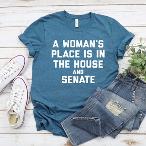 May include: A teal blue t-shirt with white text that reads "A woman's place is in the house and senate". The t-shirt is laying on a white wooden surface with a pair of white sneakers and blue jeans.