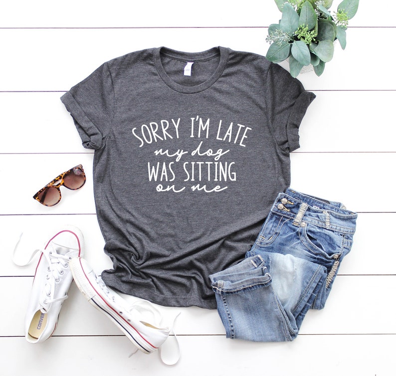 Op de afbeelding: Een grijs T-shirt met de tekst "Sorry I'm late my dog was sitting on me" in witte letters. Het shirt ligt op een wit houten oppervlak met een zonnebril, witte sneakers en een blauwe jeans.