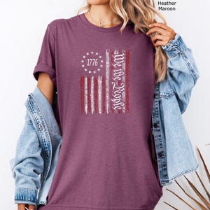 May include: Heather maroon t-shirt featuring a distressed American flag design. The flag includes the text "1776" in a circle of stars and "We the People" in a vertical column. The shirt is a solid color and has short sleeves.