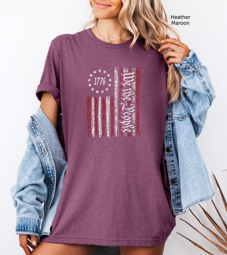 May include: Heather maroon t-shirt featuring a distressed American flag design. The flag includes the text "1776" in a circle of stars and "We the People" in a vertical column. The shirt is a solid color and has short sleeves.