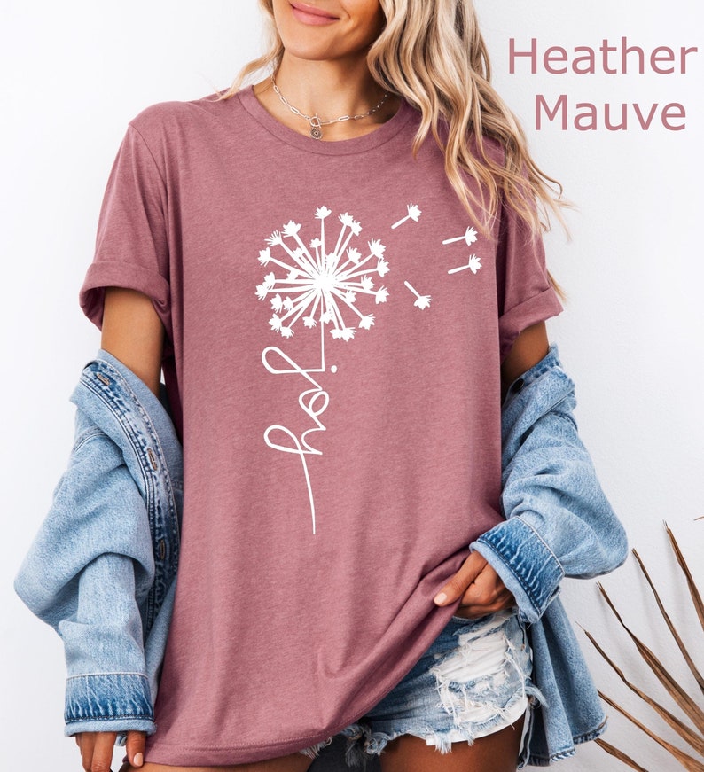 K&ouml;nnte beinhalten: Ein T-Shirt in der Farbe Heather Mauve mit einem wei&szlig;en L&ouml;wenzahnmuster und dem Wort "Hope" in Kursivschrift.
