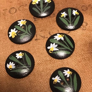 Puede incluir: Seis botones negros con margaritas pintadas de blanco y amarillo y hojas verdes.