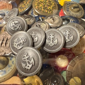 Può includere: Una collezione di bottoni color argento con un design a forma di ancora. I bottoni sono disposti in un cerchio e sono circondati da altri bottoni di vari colori e design.