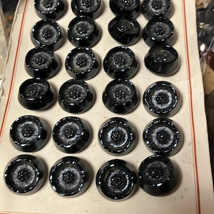 May include: A sheet of black buttons with a raised design. The buttons are arranged in rows on a white card with a red border.