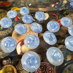 May include: A collection of 15 light blue glass buttons with a raised, textured design. The buttons are arranged in a random pattern on a surface covered in various objects.