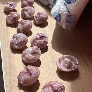 Könnte beinhalten: Eine Sammlung von 12 rosa und weißen, spiralförmigen Glasknöpfen. Die Knöpfe sind in einer Reihe auf einer Holzoberfläche angeordnet.