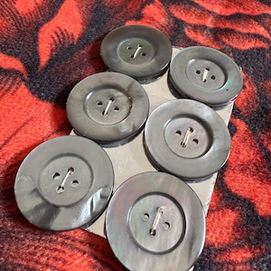 May include: Six large, round, gray buttons with a pearlescent sheen. Each button has two holes for sewing. The buttons are arranged on a small, silver metal surface, set against a red and black floral patterned background.