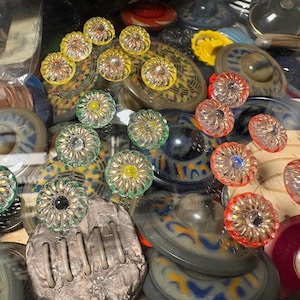 Könnte beinhalten: Eine Sammlung bunter Glas-Knöpfe und dekorativer Elemente. Die Knöpfe weisen verschiedene Designs auf, darunter blütenartige Muster mit gelben, grünen und roten Akzenten. Weitere Elemente sind runde und ovale Formen in Blau-, Braun- und Gelbtönen.