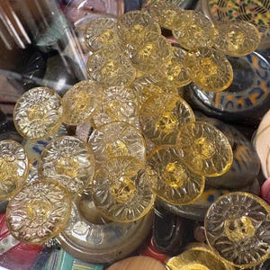 Può includere: Una collezione di bottoni vintage in vari colori e design. L'assortimento comprende bottoni trasparenti, gialli e dorati con motivi floreali e geometrici. Alcuni bottoni sono in vetro, altri sembrano essere in metallo o legno.