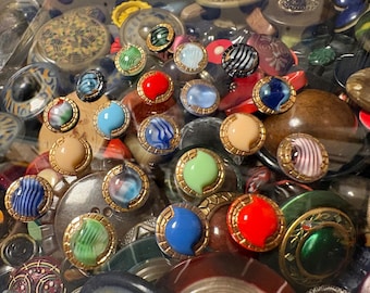 May include: A collection of assorted round buttons featuring various colors and patterns, including solid red, blue, green, and cream, alongside striped and marbled designs. Each button is framed by a metallic gold-toned textured rim.