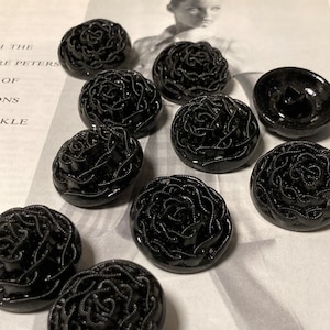 Puede incluir: Diez botones de plástico negro con un diseño de rosa. Los botones están dispuestos en semicírculo sobre una superficie blanca.