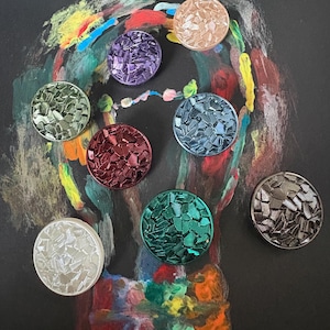 Può includere: Una collezione di otto bottoni circolari caratterizzati da una superficie testurizzata simile a un mosaico. I bottoni sono presentati in vari colori metallizzati, tra cui argento, oro, viola, verde, blu, rosso e grigio scuro, con finitura lucida.