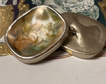 May include: Two silver-toned metal buttons, one with a pearlescent iridescent finish and the other with a smooth, matte finish.