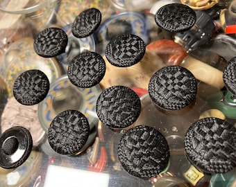 May include: A collection of black buttons with a woven pattern. The buttons are arranged on a clear surface with other objects in the background.