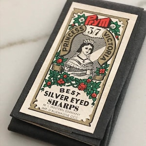 May include: A package of sewing needles labeled "Princess Victoria" with the text "Best Silver Eyed Sharps." The label features an illustration of a woman in a crown, surrounded by floral designs. The package is folded and has a dark gray color.