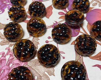 May include: A collection of 15 brown and black glass buttons with a textured, bubble-like surface. The buttons are arranged in a semi-circle on a floral patterned surface.