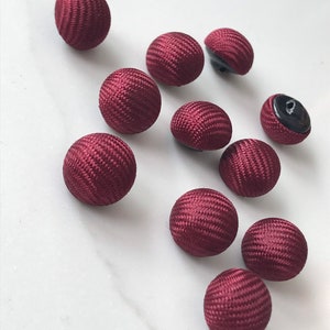 Può includere: Dieci bottoni rotondi in tessuto color borgogna con un centro nero. I bottoni sono testurizzati.