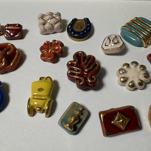 Może przedstawiać: Kolekcja różnorodnych ceramicznych guzików o rozmaitych kształtach i kolorach, w tym niebieska podkowa, żółty powóz, czerwony prezent, biały kwiat i turkusowy kwadrat ze złotymi akcentami. Każdy guzik posiada błyszczące wykończenie.