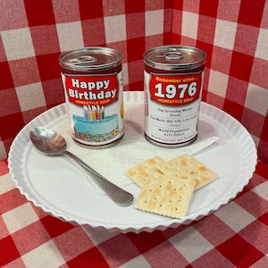 May include: Two red and white soup cans with a birthday cake and the year 1976 on them. The cans are on a white plate with two crackers. The background is a red and white checkered tablecloth.