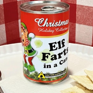 May include: A cylindrical metal can with a pull-tab lid, featuring a red and white label. The label displays an illustration of an elf, the text Christmas Holiday Collection, and the words Elf Farts in a Can, with a yellow caution warning.