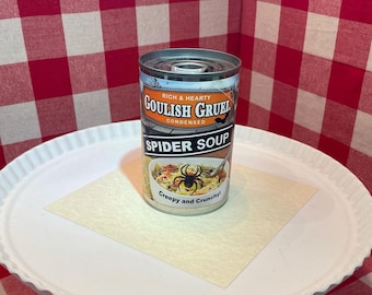 Puede incluir: Una lata de sopa de metal etiquetada como Goulish Gruel Spider Soup. La etiqueta presenta una pancarta naranja con el texto Rich & Hearty, un gráfico de araña negra y un tazón de sopa con arañas. El texto inferior dice Creepy and Crunchy!