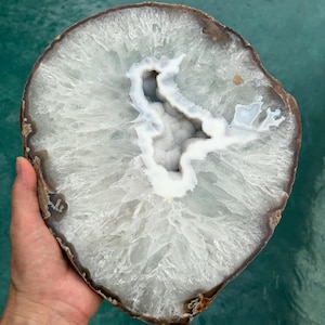 May include: A large, circular agate slice featuring a translucent, crystalline white and clear interior with a jagged, hollow center. The outer edge is defined by a thin, dark brown, earthy crust that frames the intricate, radiating mineral patterns.