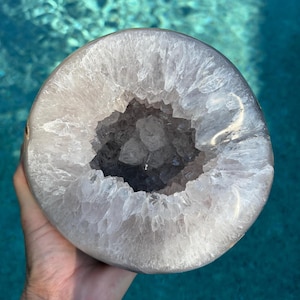 May include: A circular geode featuring a smooth, polished outer edge and a hollow center filled with jagged, translucent white and light purple quartz crystals. The mineral specimen displays a natural crystalline structure throughout.