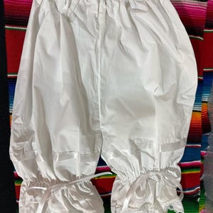 May include: White cotton bloomers with a gathered elastic waistband and lace trim. The bloomers have a satin ribbon tie at the bottom of each leg.