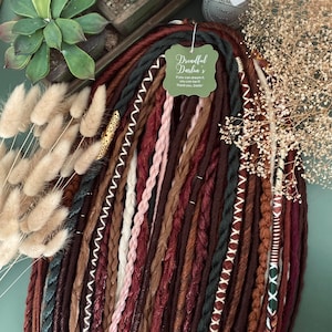 May include: A set of brown, burgundy and green synthetic dreadlocks with white and gold accents. The dreadlocks are arranged in a fan shape and have a tag that reads "Dreadful Darlin's. If you can dream it, you can be it. Thank you, Darlin."