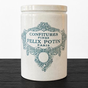 Può includere: Un barattolo di ceramica bianco con coperchio, con un'etichetta decorativa color turchese. L'etichetta recita "CONFITURES FINES FELIX POTIN PARIS" in uno stile vintage ornato. Il barattolo è cilindrico e poggia su una superficie di legno scuro.