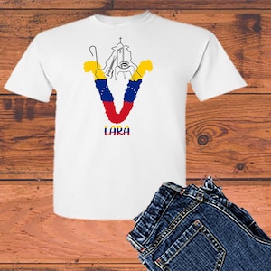 Può includere: Maglietta bianca con una grafica a forma di 'V' realizzata con i colori della bandiera venezuelana. La 'V' è composta da una chiesa con una croce in cima, un bastone da pastore e una chitarra. La parola 'LARA' è stampata sotto la grafica.