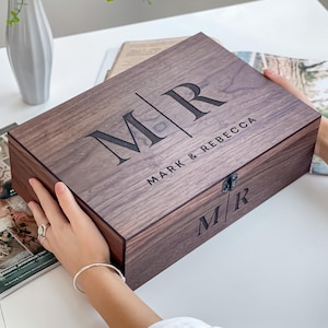 Op de afbeelding: Een rechthoekige houten kist in donkerbruin, gegraveerd met de initialen "MR" en de namen "MARK & REBECCA". De kist heeft een metalen sluiting en wordt vastgehouden.