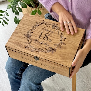 Peut inclure: Une boîte à souvenirs rectangulaire en bois avec un couvercle à charnière et un loquet en métal. Le couvercle présente une couronne de laurier gravée au laser entourant le texte "Happy 18th Birthday". La date "22-08-2027" est gravée sur le devant.
