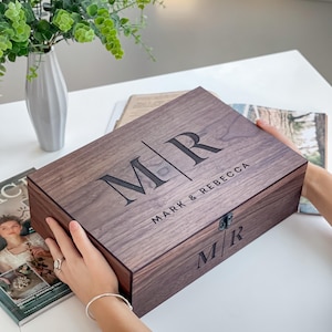 May include: A rectangular wooden box with the initials "MR" and the names "MARK & REBECCA" engraved on the lid. The box is a dark brown color and has a metal clasp. The box is being held by a person.