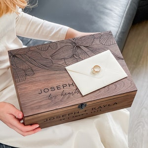 May include: A wooden keepsake box with engraved floral design and the names "JOSEPH + KAYLA" and wedding date. Two gold wedding rings rest on a cream envelope atop the box. The box reads "two hearts".