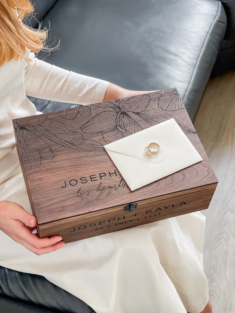 May include: A wooden keepsake box with engraved floral design and the names "JOSEPH + KAYLA" and wedding date. Two gold wedding rings rest on a cream envelope atop the box. The box reads "two hearts".