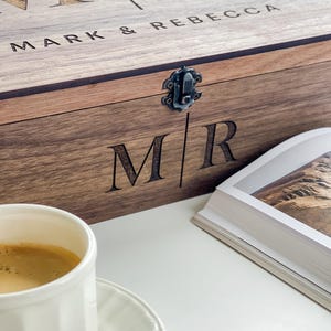 May include: A wooden box with the engraved initials "MR" and the names "MARK & REBECCA". The box has a metal clasp and is next to a cup of coffee and a book. The box is made of a light brown wood.