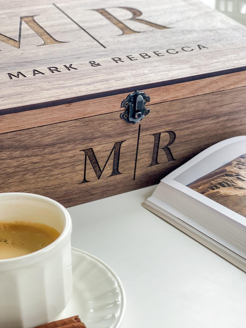 May include: A wooden box with the engraved initials "MR" and the names "MARK & REBECCA". The box has a metal clasp and is next to a cup of coffee and a book. The box is made of a light brown wood.