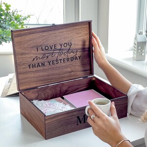May include: A wooden keepsake box with the inscription "I LOVE YOU more today THAN YESTERDAY" on the lid. The box is open, revealing a pink notebook and other items. The letter "M" is on the front of the box.