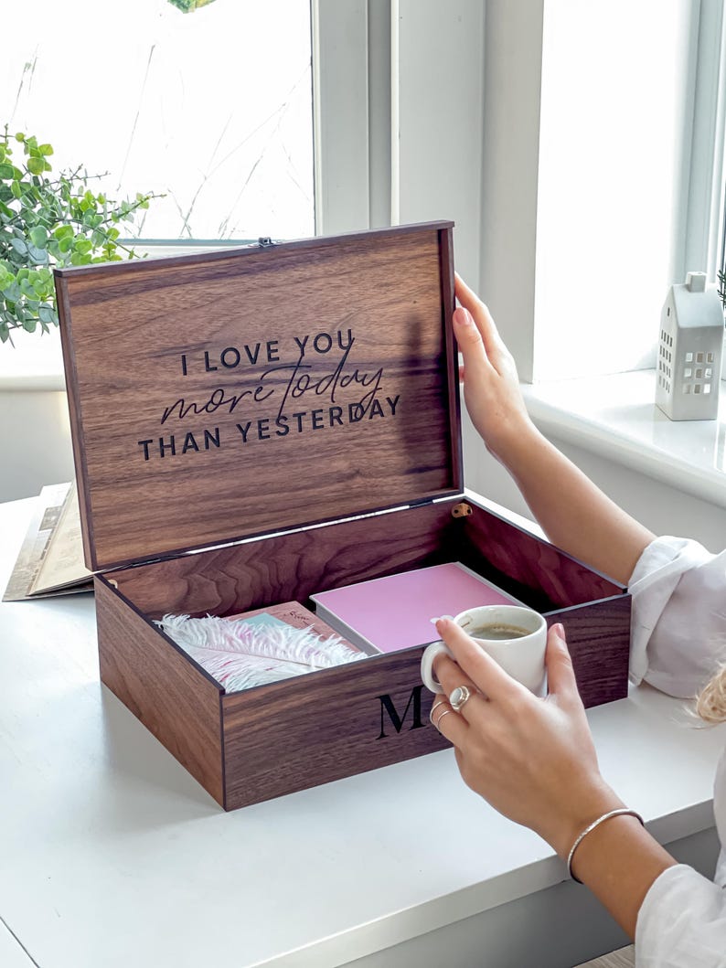 May include: A wooden keepsake box with the inscription "I LOVE YOU more today THAN YESTERDAY" on the lid. The box is open, revealing a pink notebook and other items. The letter "M" is on the front of the box.