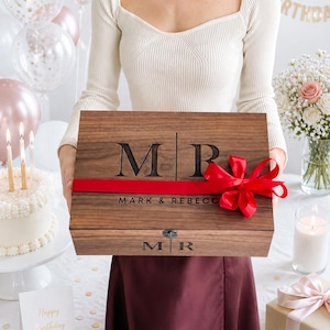 May include: A wooden gift box with the initials "MR" and "MARK & REBECCA" engraved on the lid. The box is tied with a red ribbon and bow. The background includes a birthday cake, balloons, and a "Happy Birthday" card.